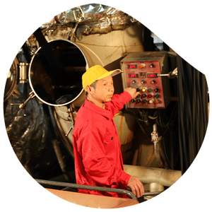 A worker wearing red overalls and a yellow cap operates an industrial control panel in a factory setting. A large metallic tank with an open hatch is visible in the background.