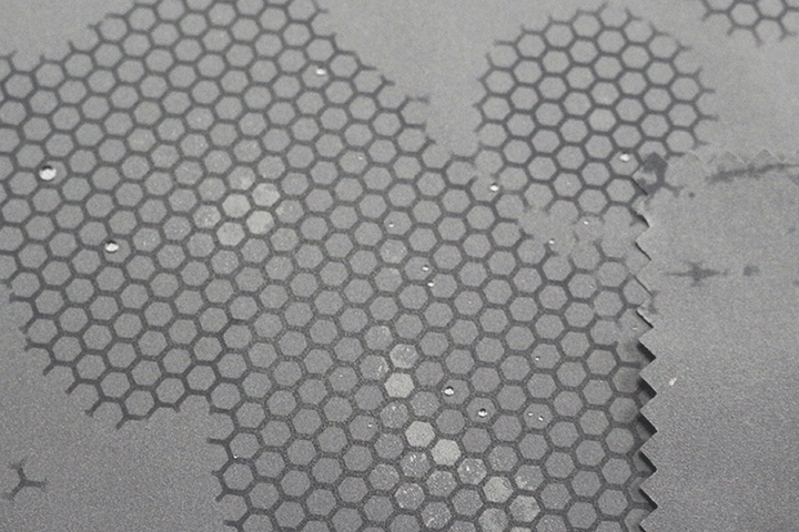 Close-up of dark grey technical fabric with a hexagonal pattern and water-repellent surface, partially covered with water droplets.
